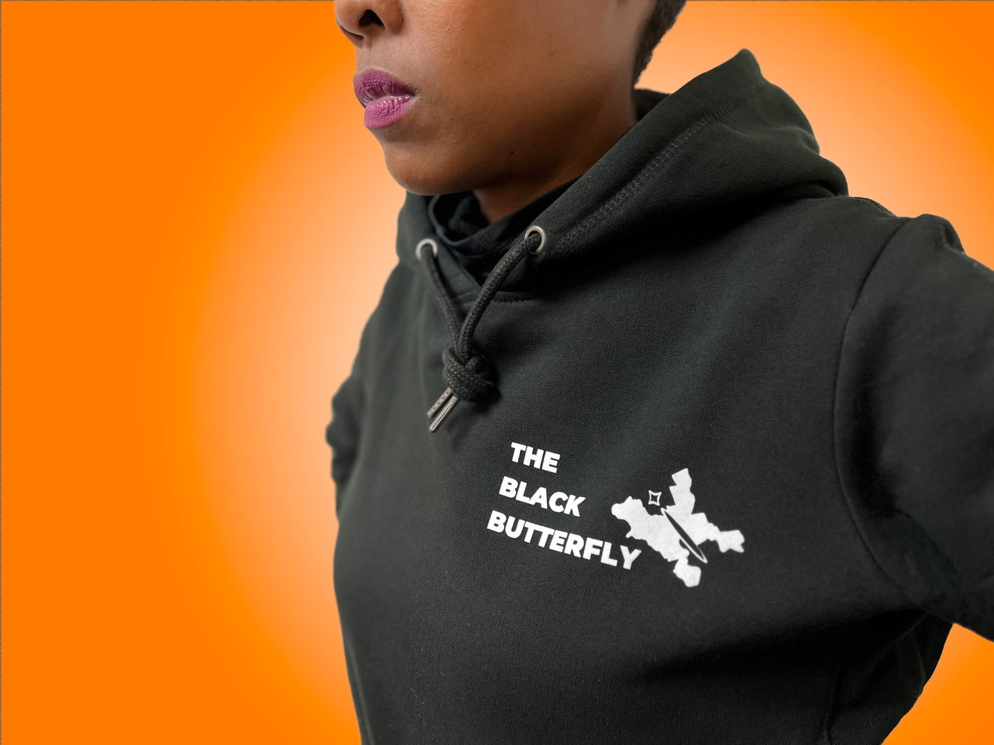 Close up of black hoodie sweatshirt with "The Black Butterfly" Lettering and butterfly design in white. Butterfly design is outline of West & East Baltimore neighborhoods on the left front chest. Worn by Black woman with magenta lipstick.