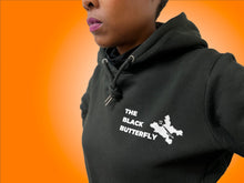 Load image into Gallery viewer, Close up of  black hoodie sweatshirt with &quot;The Black Butterfly&quot; Lettering and butterfly design in white. Butterfly design is  outline of West &amp; East Baltimore neighborhoods on the left front chest. Worn by Black woman with magenta lipstick. 
