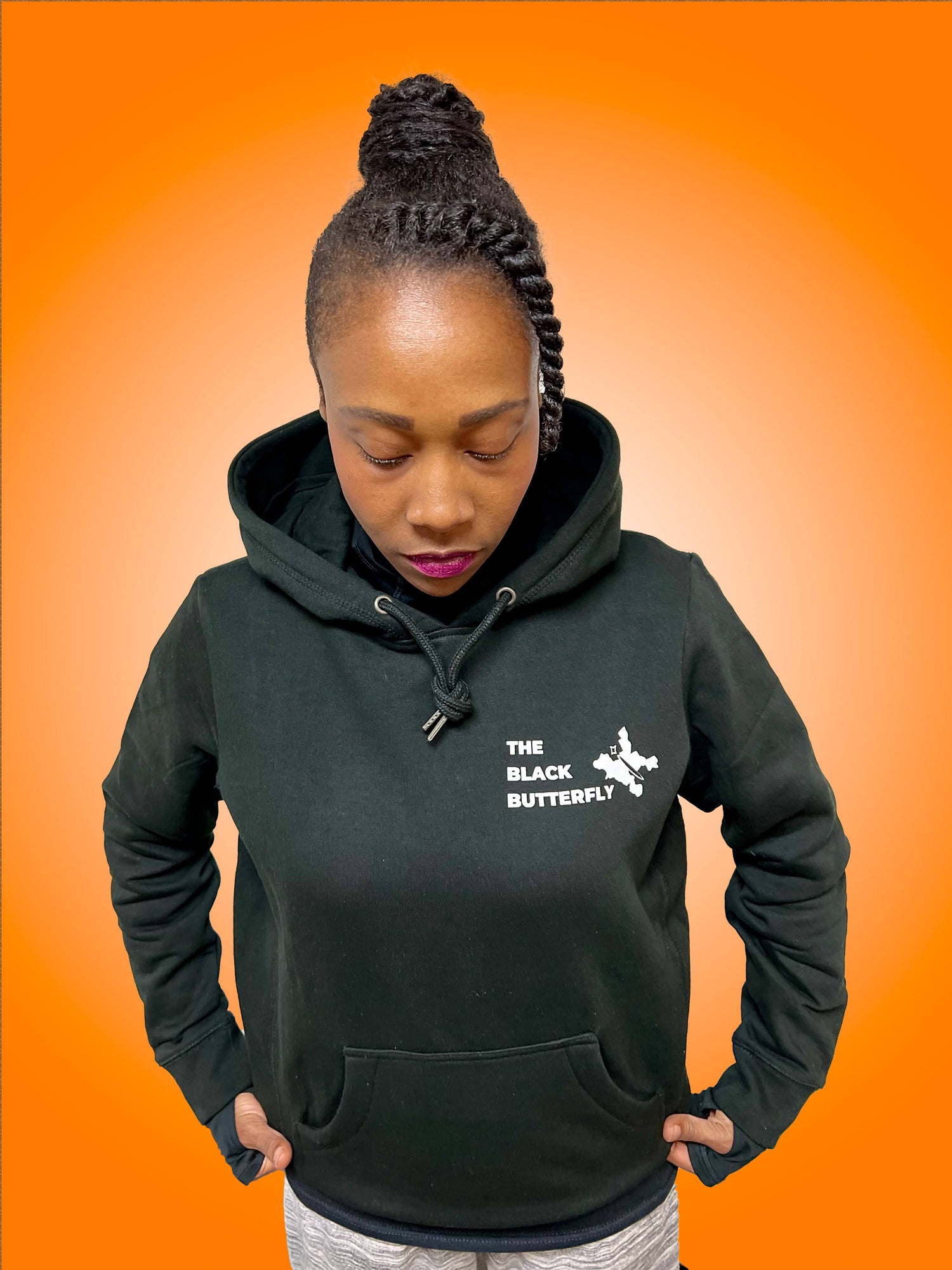 Full body shot of black hoodie sweatshirt with "The Black Butterfly" Lettering and butterfly design in white. Butterfly design is  outline of West & East Baltimore neighborhoods on the left front chest. Worn by Black woman with eyes closed, looking down, and magenta lipstick. 